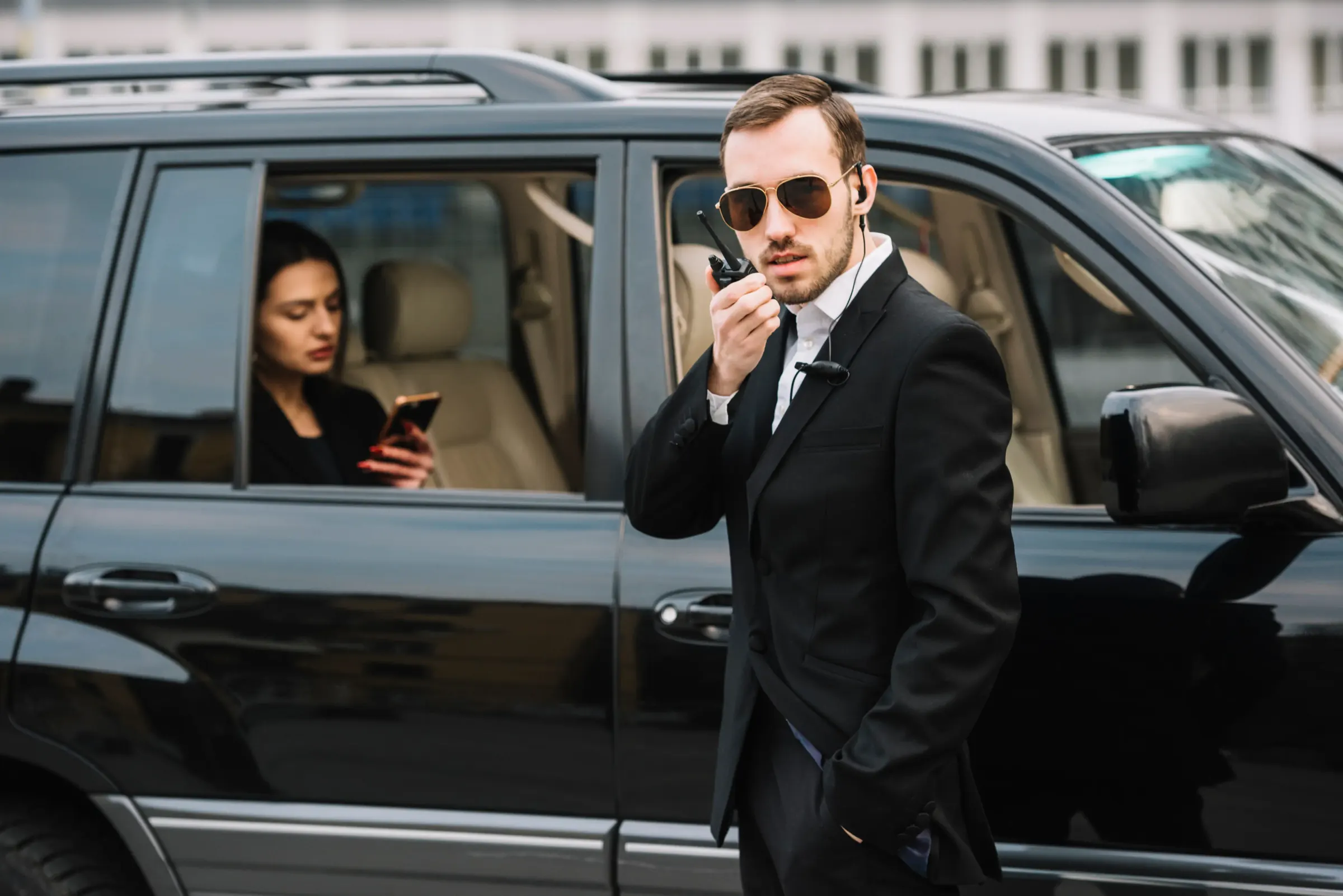 Protection agent speaking into a radio beside an SUV while the client waits inside