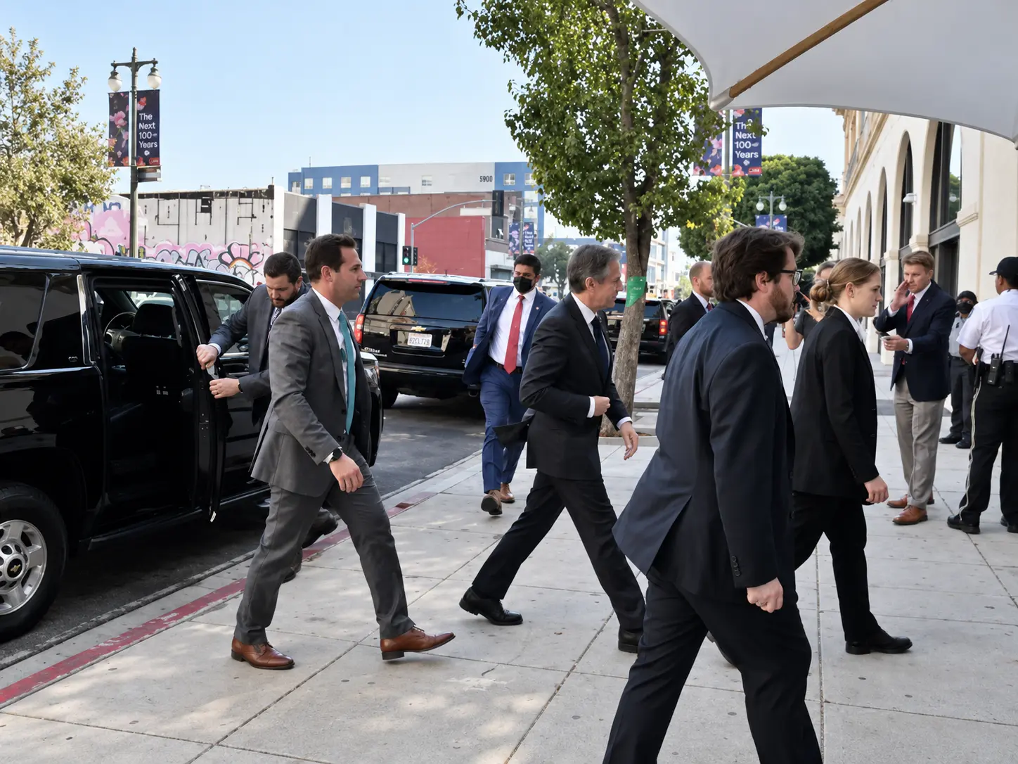 Close protection officers in suits escorting a client along a city sidewalk beside parked SUVs